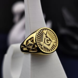 Può includere: Un anello d'oro con un simbolo massonico inciso sulla parte superiore. Il simbolo è un compasso e una squadra con un design a raggiera.