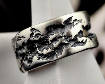May include: A wide silver-toned metal ring featuring a textured, dark, recessed pattern that wraps around the band. The surface has a polished finish on the raised areas, contrasting with the deep, jagged, and irregular black carved details.