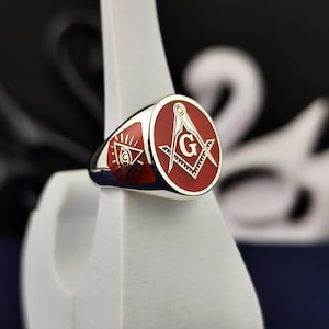 May include: A silver ring with a red enamel face featuring Masonic symbols. The ring has a polished finish and is displayed on a white stand. The symbols include a compass, square, and the all-seeing eye.