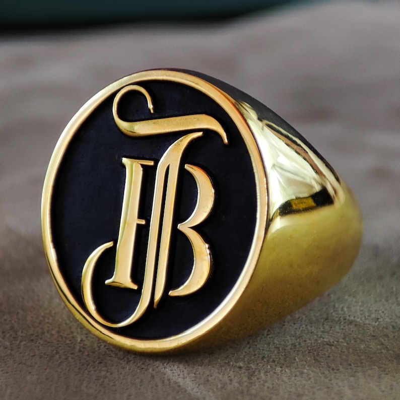 This close-up image presents a custom monogram signet ring designed with engraved initials on a smooth, polished face. The ring is available in Silver, Gold Plated, and 14k Gold variations, giving shoppers multiple material choices. The engraving is crisp and symmetrical, showing the precision of the craftsmanship. The lighting captures the reflective surface of the ring, emphasizing its luxurious appearance and suitability as a personalized Christmas gift for him or her.