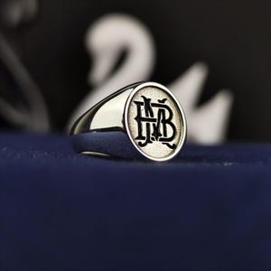 Könnte beinhalten: Ein silberner Siegelring mit einem schwarzen, gravierten Monogramm der Buchstaben "MB" auf der Oberseite.