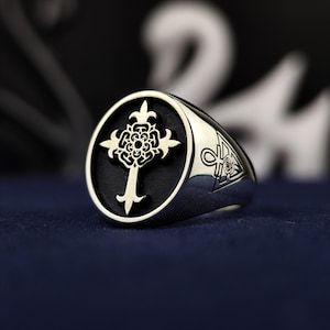 Puede incluir: Anillo de plata con un diseño de cruz detallado sobre un fondo negro. El anillo tiene una cara ovalada con un intrincado motivo de cruz y símbolos en la banda. El anillo está hecho de metal pulido.