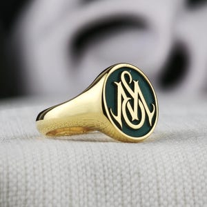 Könnte beinhalten: Ein goldener Siegelring mit einer runden, dunkelgrünen Einlage. In der Mitte der Einlage befindet sich ein erhabenes, poliertes Goldmonogramm aus den ineinander verschlungenen Buchstaben M und S in dekorativem Schreibschriftstil.