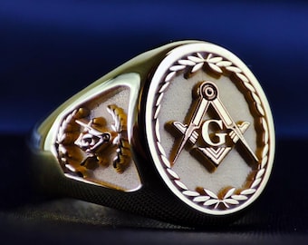 Puede incluir: Un anillo de tono dorado con una cara circular que presenta un símbolo masónico en relieve de escuadra y compás centrado con la letra G, rodeado por un borde de corona de laurel. El lateral del anillo muestra un emblema más pequeño.