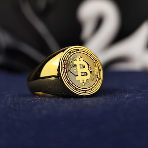 Könnte beinhalten: Ein goldener Ring mit einem Bitcoin-Symbol auf der Oberseite. Der Ring ist mit den Worten "Bitcoin Decentralized Digital Currency" und "Ultimate Digital Liberty" graviert.