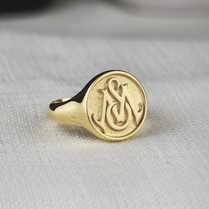 May include: A gold signet ring featuring a circular face with an engraved monogram of the letters M and S intertwined. The ring has a polished, reflective finish on the band and a textured, matte appearance within the circular face design.