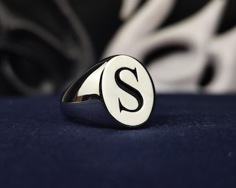 May include: A polished silver-tone signet ring featuring a smooth, flat, oval-shaped face. The center of the ring face is engraved with a large, bold, black capital letter S. The band of the ring is solid and tapers toward the back of the piece.