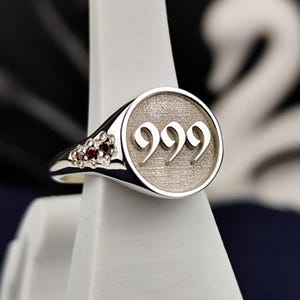 May include: A silver signet ring with a round face featuring the number 999 engraved in a bold font. The ring has a band adorned with small red gemstones.