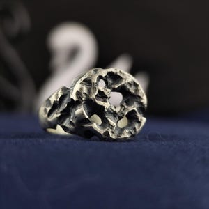 May include: A silver-toned ring featuring a highly textured, irregular surface with deep, crater-like indentations and a dark, oxidized finish. The band has a rugged, organic appearance with uneven edges and a porous, pitted metal structure.