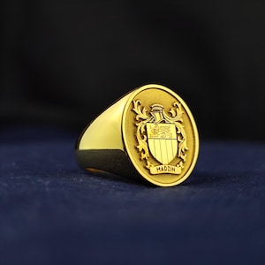 May include: A gold signet ring with an oval face featuring a detailed family crest. The crest includes a shield, helmet, and decorative flourishes. The word "MADZIN" is inscribed at the bottom of the crest. The ring is set against a dark blue background.