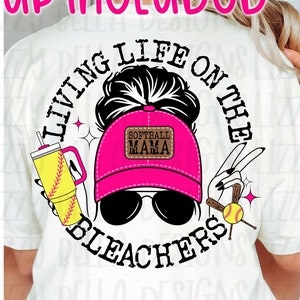 May include: A white t-shirt mockup with a graphic design featuring a woman with a bun in her hair wearing a pink baseball cap with the text "Softball Mama" on it. The graphic also includes a baseball bat, a baseball, and a drink with a straw. The text "Living Life on the Bleachers" is written in a circle around the graphic.