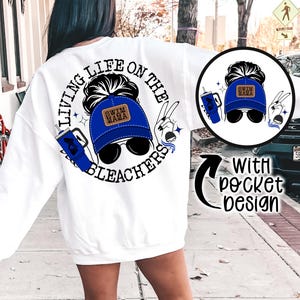 May include: White sweatshirt with a graphic design on the back that says "LIVING LIFE ON THE BLEACHERS". The design features a cartoon image of a person wearing a blue hat that says "SWIM MAMA", sunglasses, and a beverage. The sweatshirt has a pocket design.
