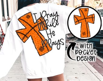 May include: White sweatshirt with a basketball-shaped cross design and the text "I Pray While He Plays" printed on the back. The sweatshirt has a pocket on the front.