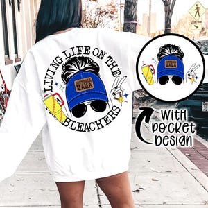 May include: White sweatshirt with a graphic print of a woman wearing a blue baseball cap with the text "Softball Mama" on it. The graphic also includes a baseball bat, a drink, and the text "Living Life on the Bleachers". The sweatshirt has a pocket on the front with a smaller version of the graphic.