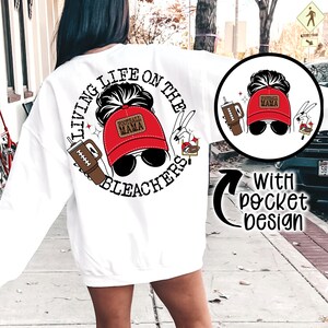 May include: White sweatshirt with a graphic print of a woman wearing a red baseball cap with the text "Football Mama" and the text "Living Life on the Bleachers".