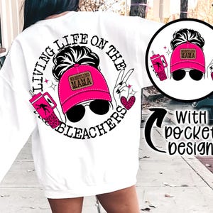 May include: White sweatshirt with a pink and black graphic design that says "Living Life on the Bleachers" with a woman's hair in a bun wearing a pink hat that says "Wrestling Mama". The sweatshirt has a pocket on the front.