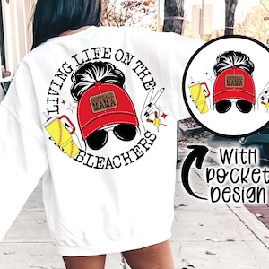 May include: White sweatshirt with a graphic design featuring a woman wearing sunglasses and a red baseball cap that says "Softball Mama." The text around the image says "Living Life on the Bleachers."