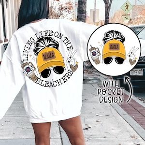 May include: White sweatshirt with a graphic print of a woman wearing a yellow baseball cap with the word "Mama" on it. The graphic also includes a drink with a straw and the text "Living Life on the Bleachers". The sweatshirt has a pocket on the front.