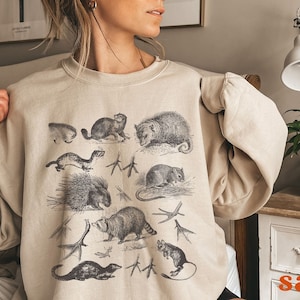 May include: A beige sweatshirt featuring a vintage illustration of various animals, including a raccoon, a porcupine, a weasel, and an opossum. The animals are printed in black and white on a light beige background.
