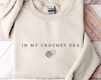 May include: Sand-colored sweatshirt with the text "IN MY CROCHET ERA" in black. A graphic of yarn and hearts is below the text. The sweatshirt is folded on a wooden surface.