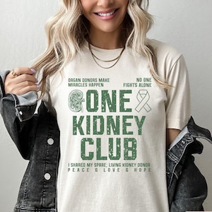 May include: A beige t-shirt with a green graphic that says "Gone One Kidney Club". The graphic also includes a kidney and a ribbon. The text below the graphic says "I shared my spare; Living Kidney Donor Peace & Love & Hope".