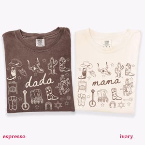 May include: Two folded t-shirts, one espresso and one ivory, with cowboy-themed illustrations. The espresso shirt says "dada" and the ivory shirt says "mama". Both shirts feature cowboy boots, hats, and other western icons.