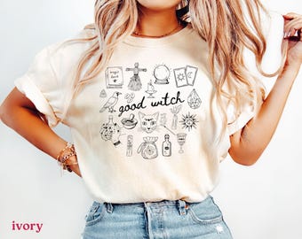 May include: Ivory-colored t-shirt featuring a black and white illustration of witchy symbols, including a cat face, crystal ball, and a book. The words "good witch" are written in a handwritten style. The shirt is a casual style.