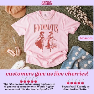 May include: Pink t-shirt featuring a vintage illustration of two women in red dresses with the text "ROOMMATES" printed in black. The t-shirt is laid flat on a brown surface with a hat, hair clips, and a blanket with hearts.