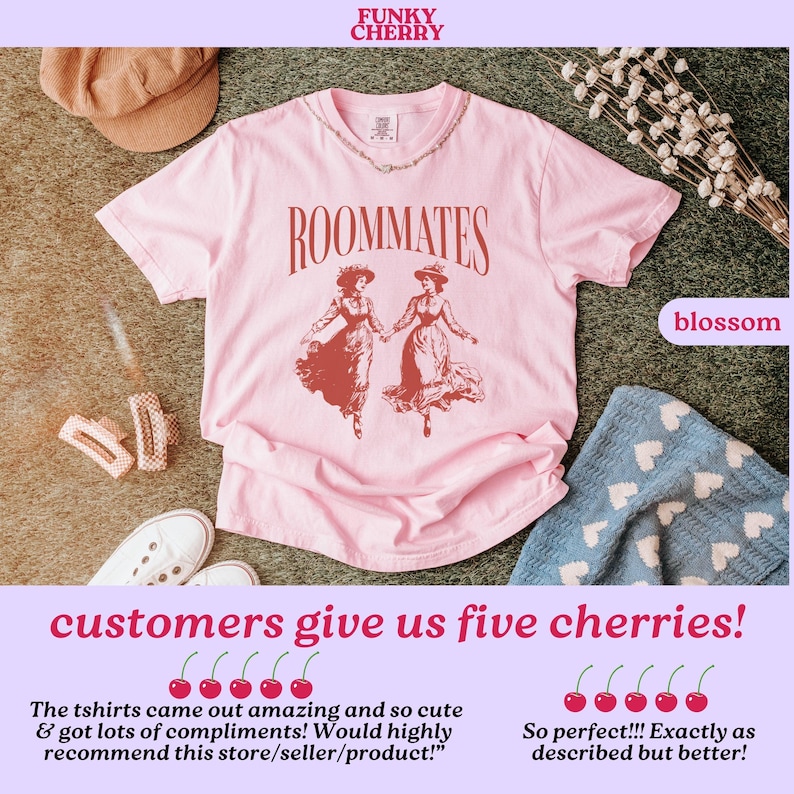 May include: Pink t-shirt featuring a vintage illustration of two women in red dresses with the text "ROOMMATES" printed in black. The t-shirt is laid flat on a brown surface with a hat, hair clips, and a blanket with hearts.