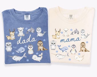 Puede incluir: Dos camisetas Comfort Colors, una azul vaquero y otra marfil. Cada camiseta presenta una colección de criaturas marinas y animales del bosque ilustrados. La camisa azul dice "dada" y la camisa marfil dice "mama".