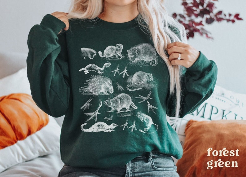 May include: A forest green sweatshirt featuring a white print of various woodland animals, including a raccoon, a porcupine, a possum, and a squirrel.
