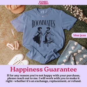 May include: A blue t-shirt with the text "ROOMMATES" printed on the front. The shirt features a vintage illustration of two women in dresses. The shirt is laid out on a patterned surface with a hat and a pair of white shoes.