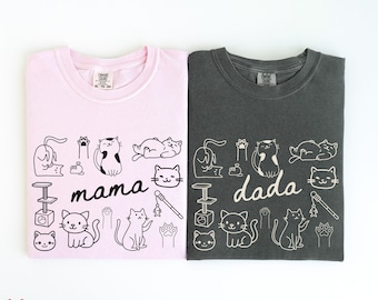 Pode incluir: Duas camisetas dobradas, uma rosa claro e outra cinza escuro. A camiseta rosa tem a palavra "mama" e a camiseta cinza tem a palavra "dada". Ambas as camisetas apresentam um padrão repetido de ilustrações de gatos e objetos relacionados a gatos.