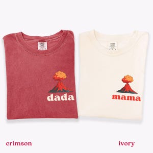 Puede incluir: Se muestran dos camisetas, carmesí y marfil. Cada camiseta presenta un gráfico de volcán con las palabras "dada" y "mama" en blanco y rojo, respectivamente. Las camisetas están hechas de un material suave.
