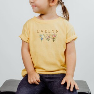 May include: Yellow t-shirt with the name "EVELYN" in brown letters above three floral bouquets. The shirt is a heather yellow gold color and is worn by a child.