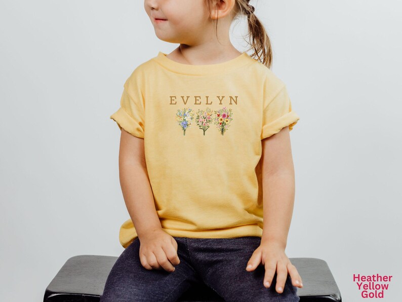 May include: Yellow t-shirt with the name "EVELYN" in brown letters above three floral bouquets. The shirt is a heather yellow gold color and is worn by a child.