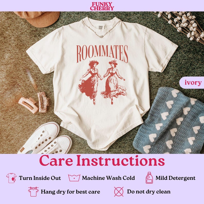 May include: A white t-shirt with the text "ROOMMATES" printed in black. The shirt features a red illustration of two women in vintage dresses. The shirt is laid out on a green surface with a blue blanket and a brown hat.
