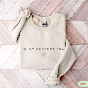 Puede incluir: Una sudadera de color arena con el texto "IN MY CROCHET ERA" y un peque&ntilde;o gr&aacute;fico de un coraz&oacute;n con hilo y un gancho de ganchillo.