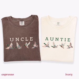 Peut inclure: Deux t-shirts, un espresso et un ivoire, chacun avec un col rond. Le t-shirt espresso porte le mot "UNCLE" au-dessus d'une rang&eacute;e de canards en vol. Le t-shirt ivoire porte le mot "AUNTIE" au-dessus d'une rang&eacute;e de canards en vol.