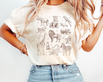 May include: Ivory-colored t-shirt featuring a collection of safari animals in a vintage style. The design includes an elephant, leopard, parrot, monkey, lion, giraffe, zebra, and more. The shirt is a relaxed fit.
