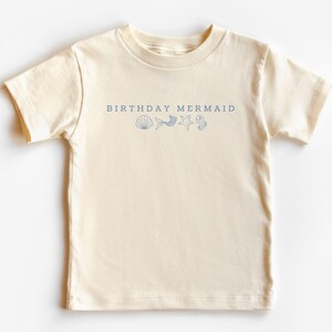 Puede incluir: Una camiseta de color natural con el texto "BIRTHAY MERMAID" en azul marino, junto con ilustraciones de conchas, cola de sirena, estrella de mar e hipocampo. La camiseta es de material suave y es adecuada para ni&ntilde;os.