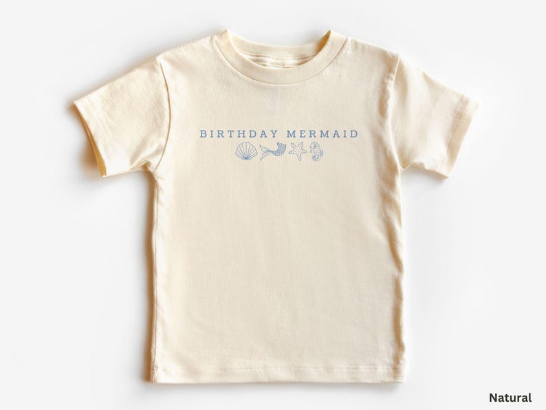 Puede incluir: Una camiseta de color natural con el texto "BIRTHAY MERMAID" en azul marino, junto con ilustraciones de conchas, cola de sirena, estrella de mar e hipocampo. La camiseta es de material suave y es adecuada para ni&ntilde;os.