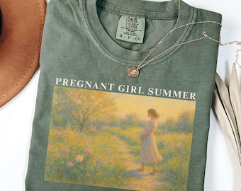May include: Moss green t-shirt with the text "PREGNANT GIRL SUMMER" above a painting of a pregnant person walking on a path. The shirt has a crew neck and short sleeves. A gold necklace with a pendant is on the shirt.