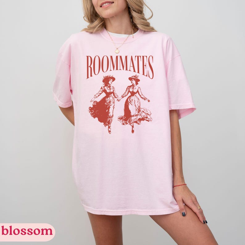 May include: A pink t-shirt with the text "ROOMMATES" printed in black. The shirt features a red illustration of two women in dresses holding hands and dancing.