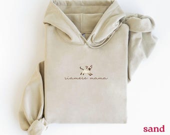 May include: A sand-colored hoodie featuring a Siamese cat graphic and the text "siamese mama" in a cursive font. The hoodie has a drawstring hood and a kangaroo pocket. The word "sand" is printed in the bottom right corner.