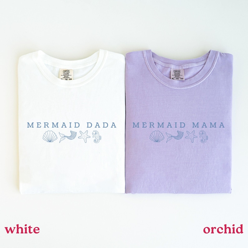 Puede incluir: Dos camisetas dobladas, una blanca y otra orqu&iacute;dea. La camiseta blanca dice "MERMAID DADA" y la camiseta orqu&iacute;dea dice "MERMAID MAMA" en texto azul. Ambas camisetas tienen una concha, una cola de sirena, una estrella de mar y un gr&aacute;fico de caballito de mar.