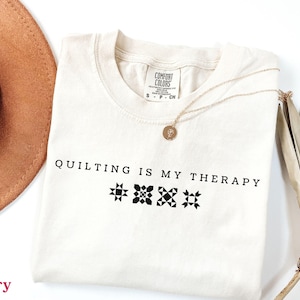 May include: Ivory-colored t-shirt with the text "QUILTING IS MY THERAPY" in black. The shirt features three quilt block designs. A gold necklace and a brown hat are also visible.