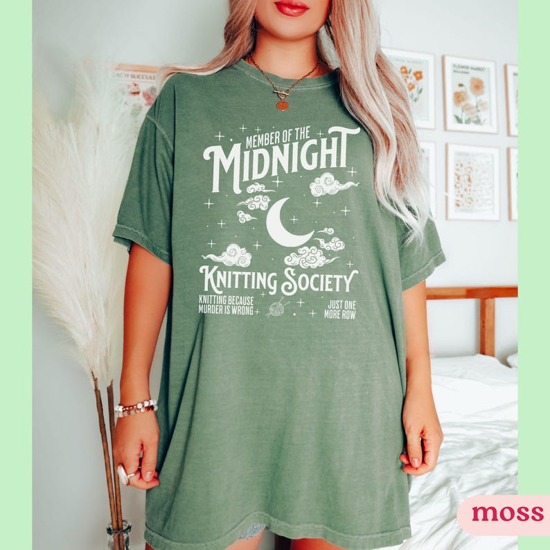 May include: A moss green t-shirt with the text "MEMBER OF THE MIDNIGHT KNITTING SOCIETY" in white. The design includes a crescent moon, clouds, and stars. The shirt also has the phrases "KNITTING BECAUSE MURDER IS WRONG" and "JUST ONE MORE ROW".