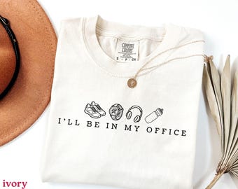 May include: Ivory-colored t-shirt with the text "I'll Be In My Office" and line drawings of sneakers, a watch, headphones, and a water bottle. A gold necklace and a brown hat are also visible.