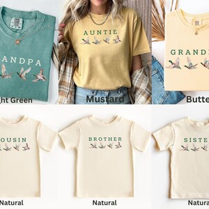 Peut inclure: Plusieurs t-shirts de couleurs vert clair, moutarde et naturel. Chaque t-shirt affiche un titre familial tel que "Grand-p&egrave;re" ou "Tante" en vert, accompagn&eacute; d'un motif de canard.